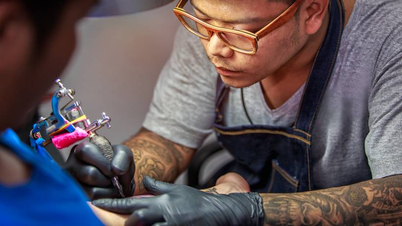 A male tattoo artist with glasses and a denim apron focuses on inking a client’s forearm using a traditional coil machine. - earn money as a tattoo artist