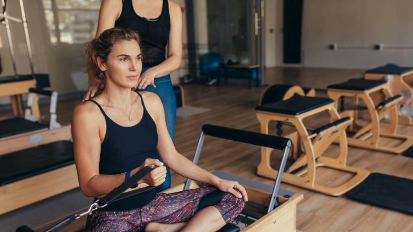 A woman sits on a Pilates training machine reformer and uses a hand strap while receiving guidance from a teacher. - how much does pilates class cost