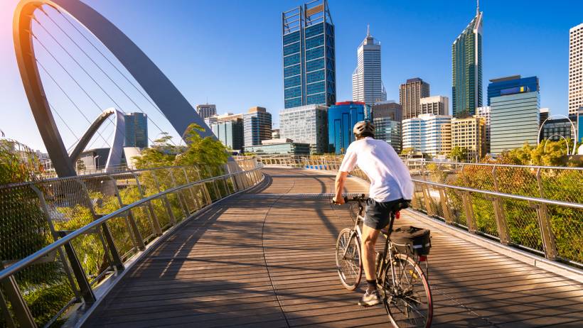 A cyclist in a white shirt and helmet cycling on Elizabeth Quay Bridge with the Perth city skyline and the Spanda sculpture in the background under a clear blue sky. - moving to perth