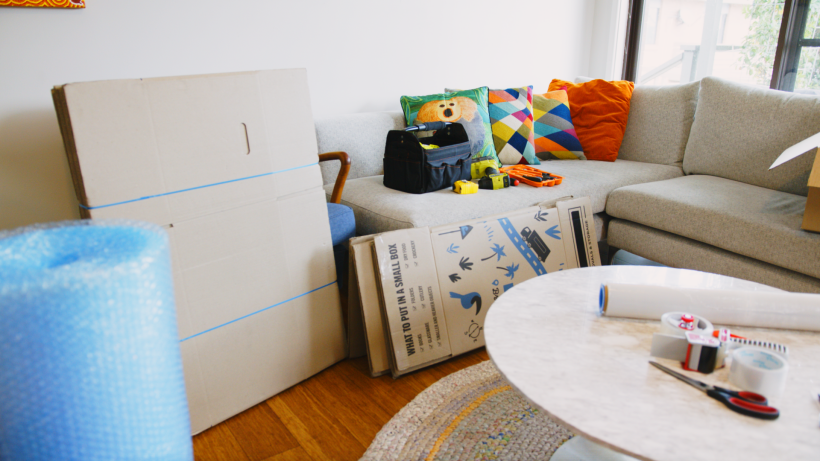 Living room filled with supplies for planning moving, including flat-packed cardboard boxes, bubble wrap, packing tape, and a tool bag on a grey sofa. - move to perth