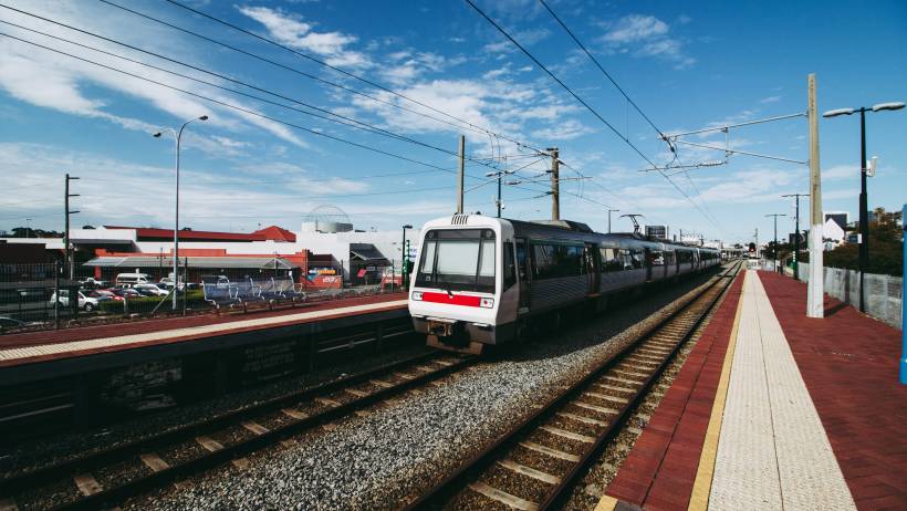 A silver and white Perth train pulled into a station platform under a bright blue sky with suburban buildings in the background. - perth relocation