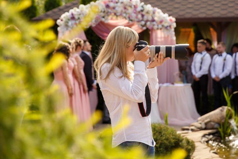 how to make money with photography - Wedding photographer taking pictures at an outdoor ceremony with the bridal party in the background