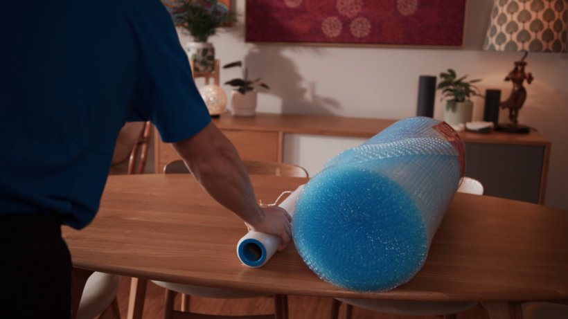 Open cardboard box with packing supplies on a wooden table. - how to pack glasses for movingA person in a blue shirt placing a large roll of blue bubble wrap and white foam packing material on a wooden dining table. - how to pack glasses when moving