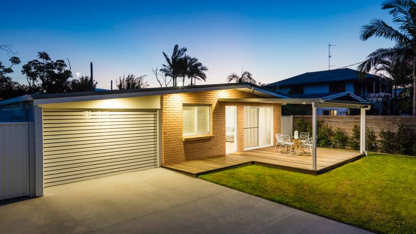 Single-story brick house at dusk with an illuminated garage, manicured lawn, and a wooden outdoor deck. - moving to gold coast