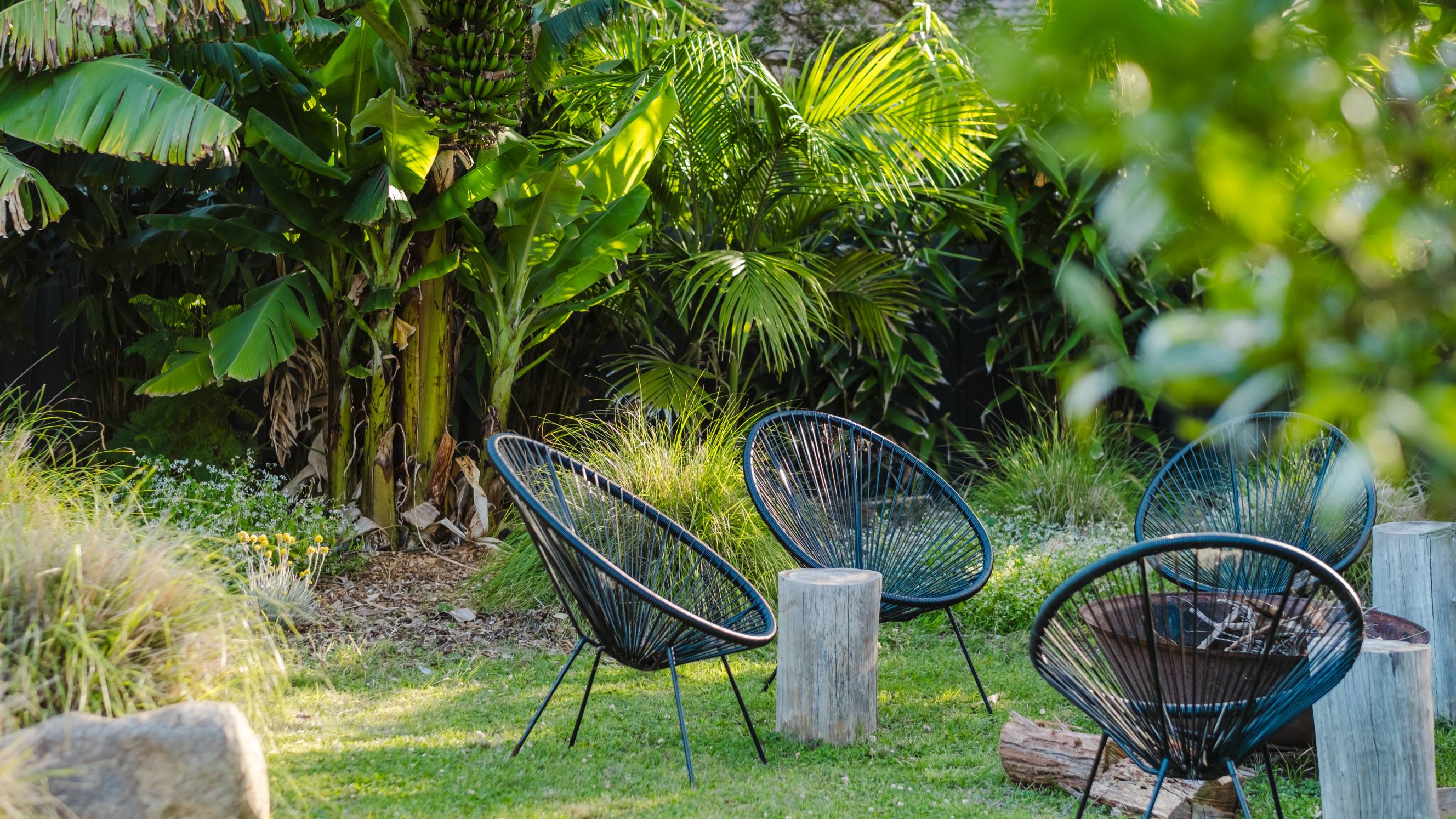 An outdoor seating area with string chairs and rustic wooden log side tables situated in a lush tropical backyard.