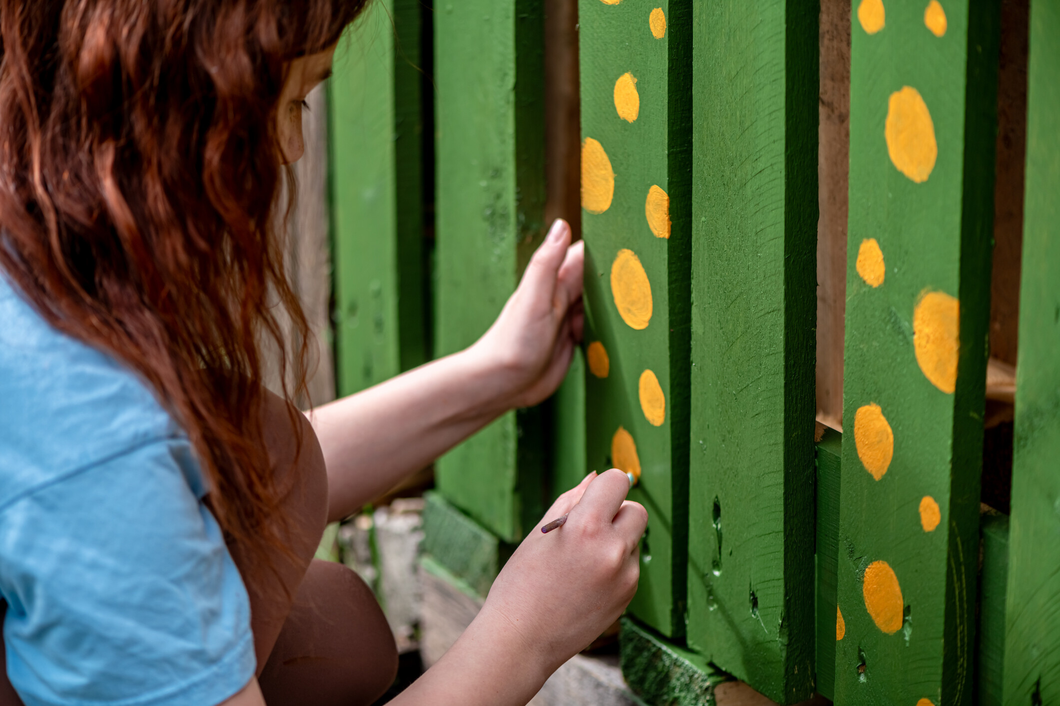 A close-up of a woman adding unique yellow patterns to a green timber fence. - garden fencing ideas