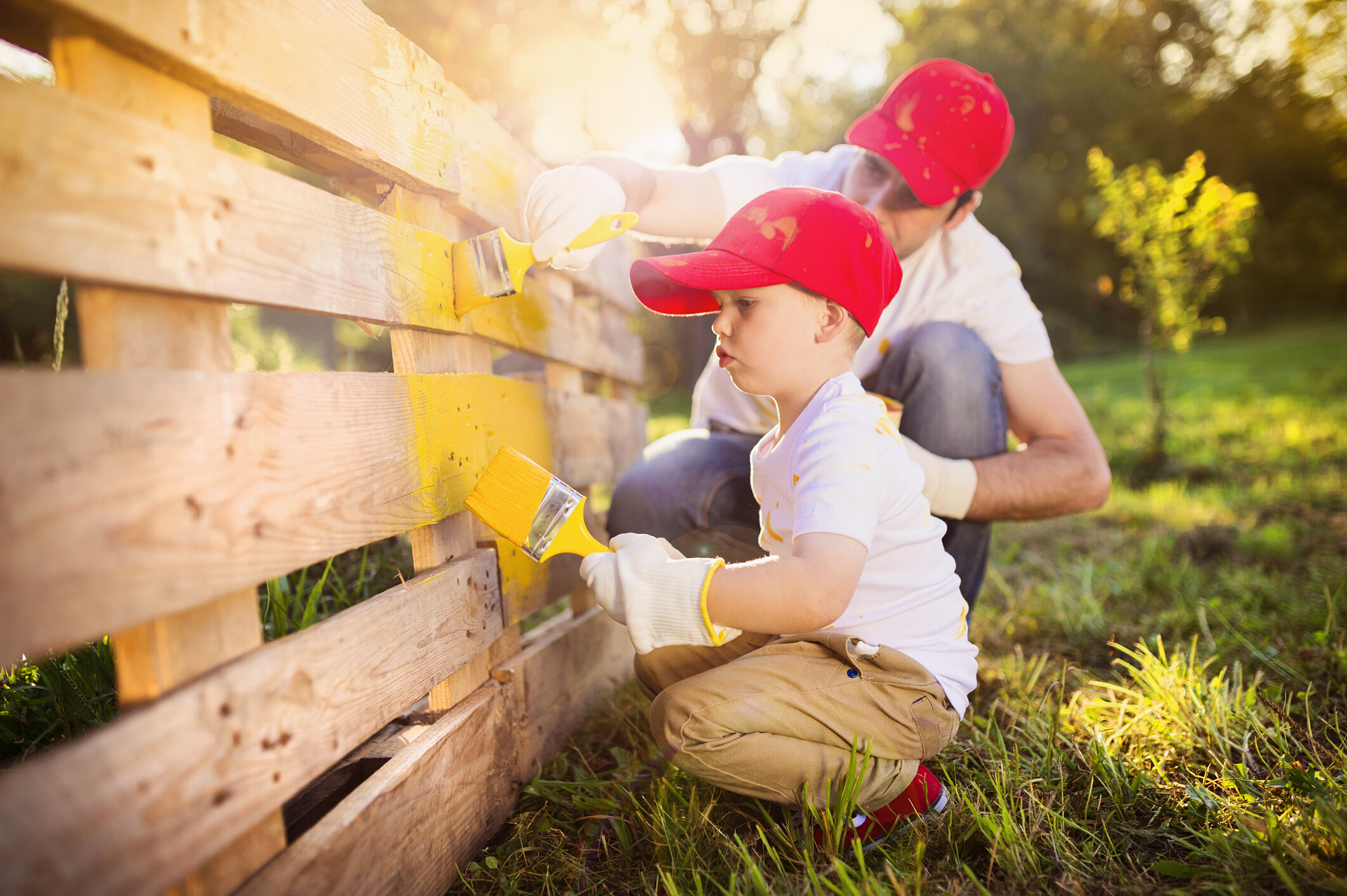 A father and young child using paintbrushes to paint a wooden fence yellow. - garden fencing ideas