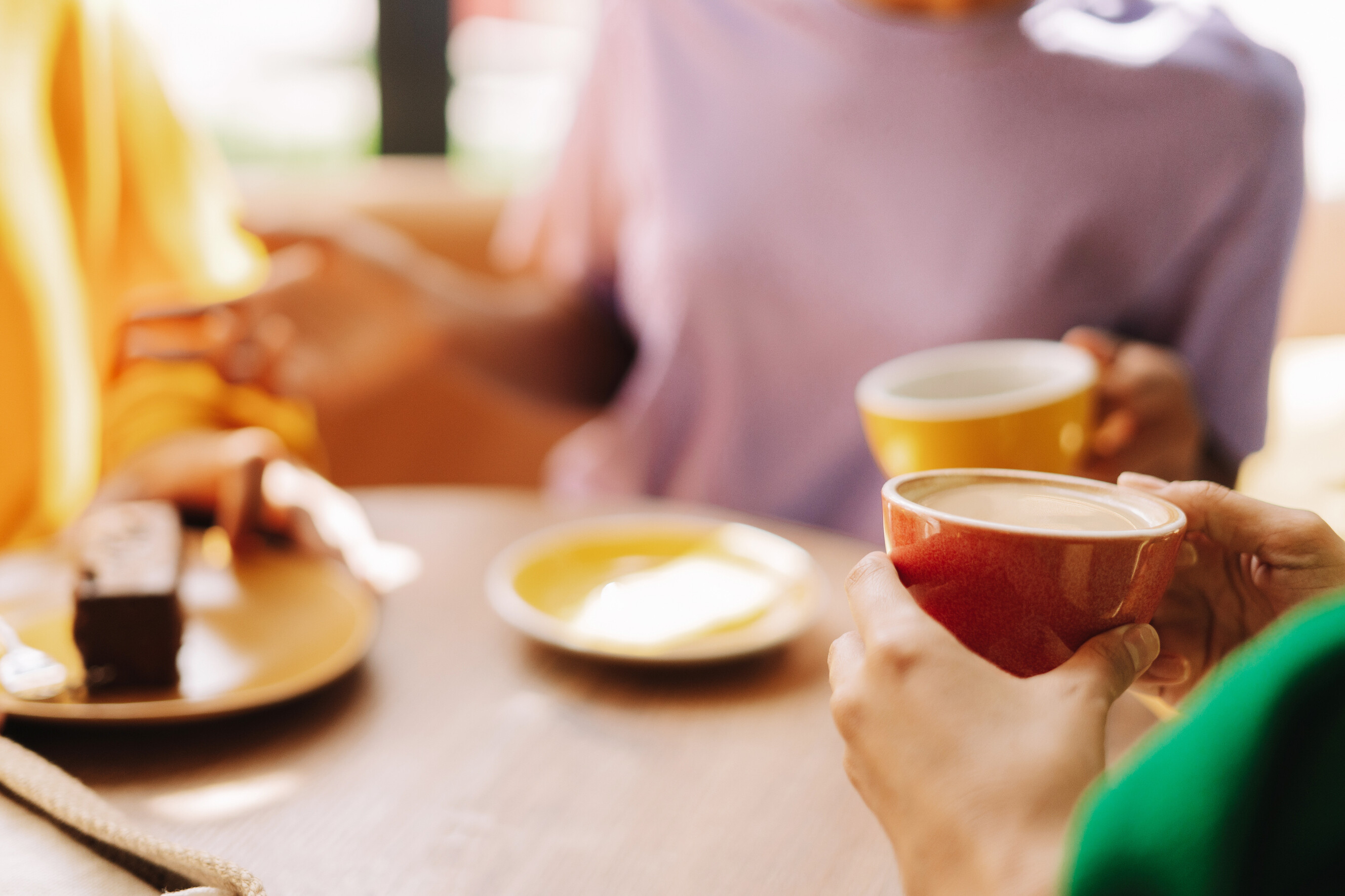Hands holding an orange and yellow coffee cup while chatting over cake.