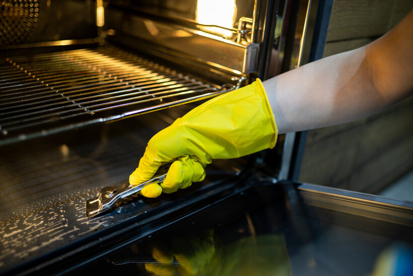A gloved hand using a scraper to remove grease and grime from an oven. - café tasks