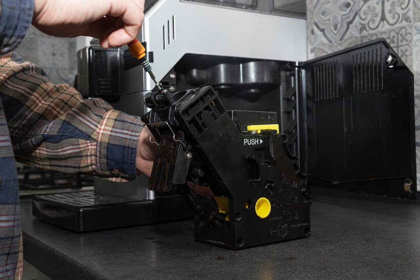 A man using an orange-handled screwdriver to open a coffee machine’s brew unit. - café tasks