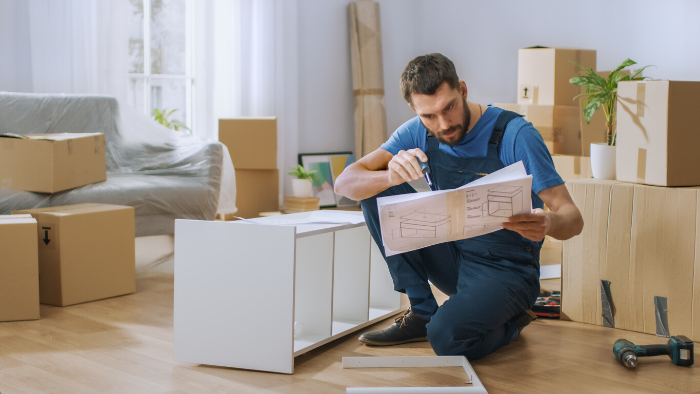 Furniture assembler checking instructions while building a cabinet in a room filled with moving boxes.