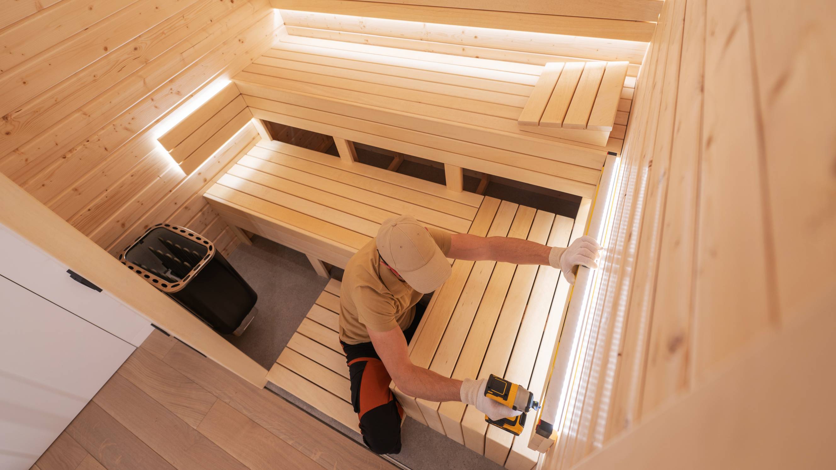 A technician using a power drill to assemble wooden seating during a custom sauna installation.