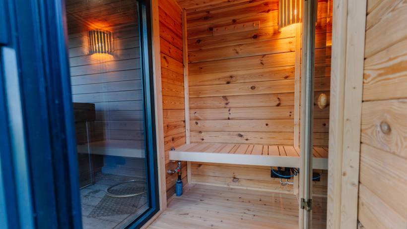 A small wooden sauna interior featuring light timber paneling, slatted bench seating, and warm sconce lighting. - sauna installation cost