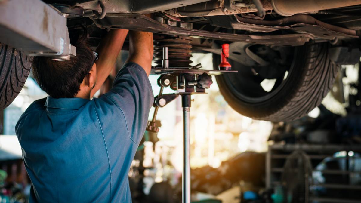 A mechanic works beneath a raised vehicle, carefully removing or replacing a catalytic converter using a transmission jack and hand tools.