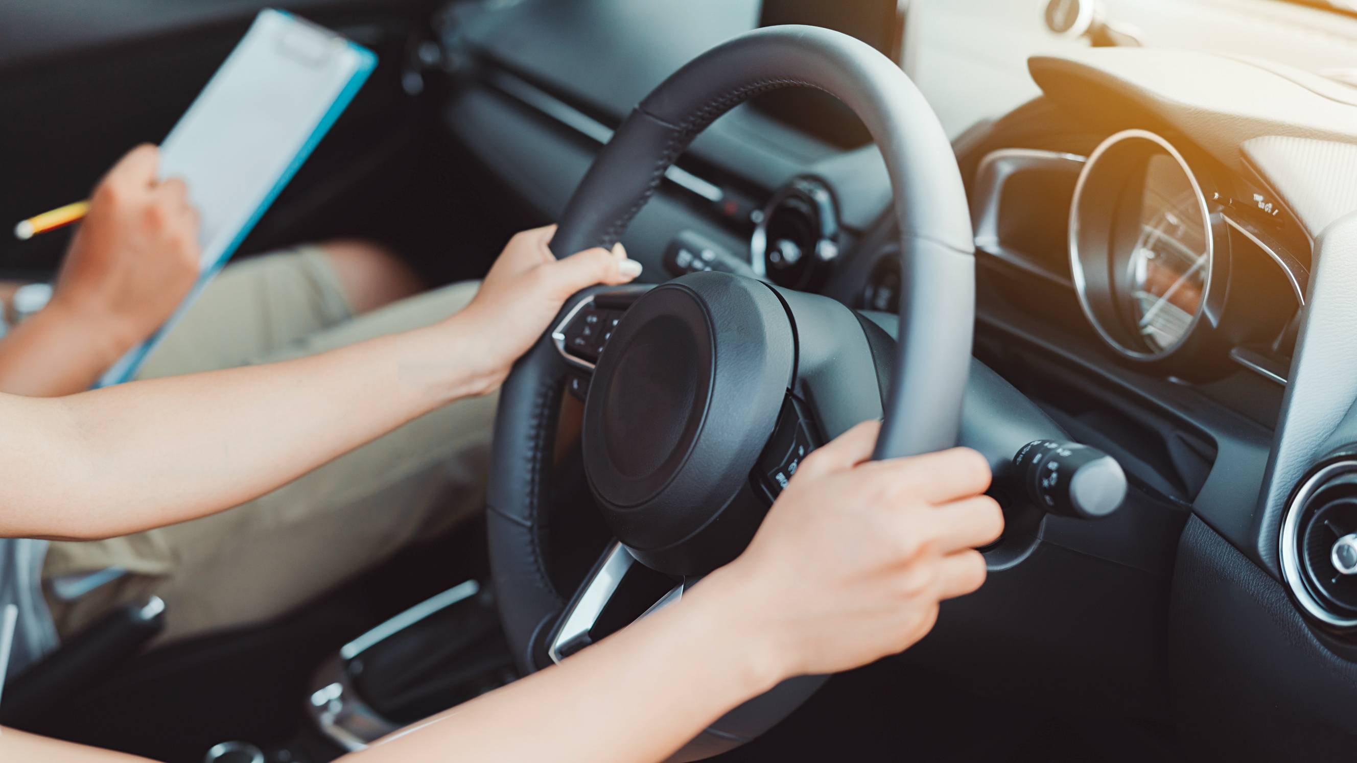 A person gripping a steering wheel during a driving lesson, with an instructor holding a clipboard