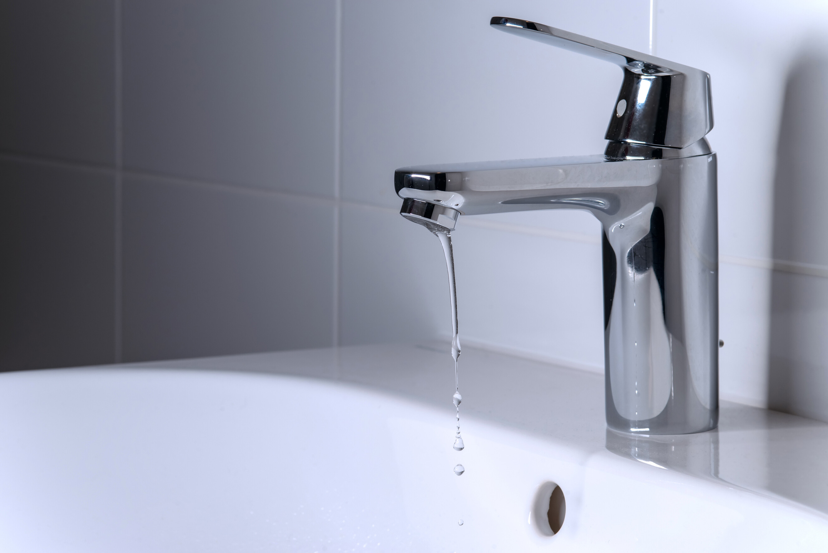 A leaking chrome bathroom tap with a slow drip falling into a white porcelain basin.