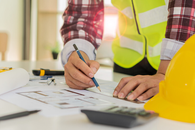 Hands sketch on building plans beside a yellow hard hat and tools. - brick garage cost