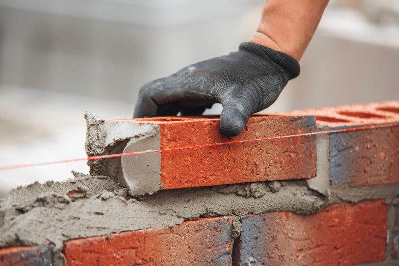 Gloved hands lay red brick over a bed of wet mortar during construction. - brick garage cost