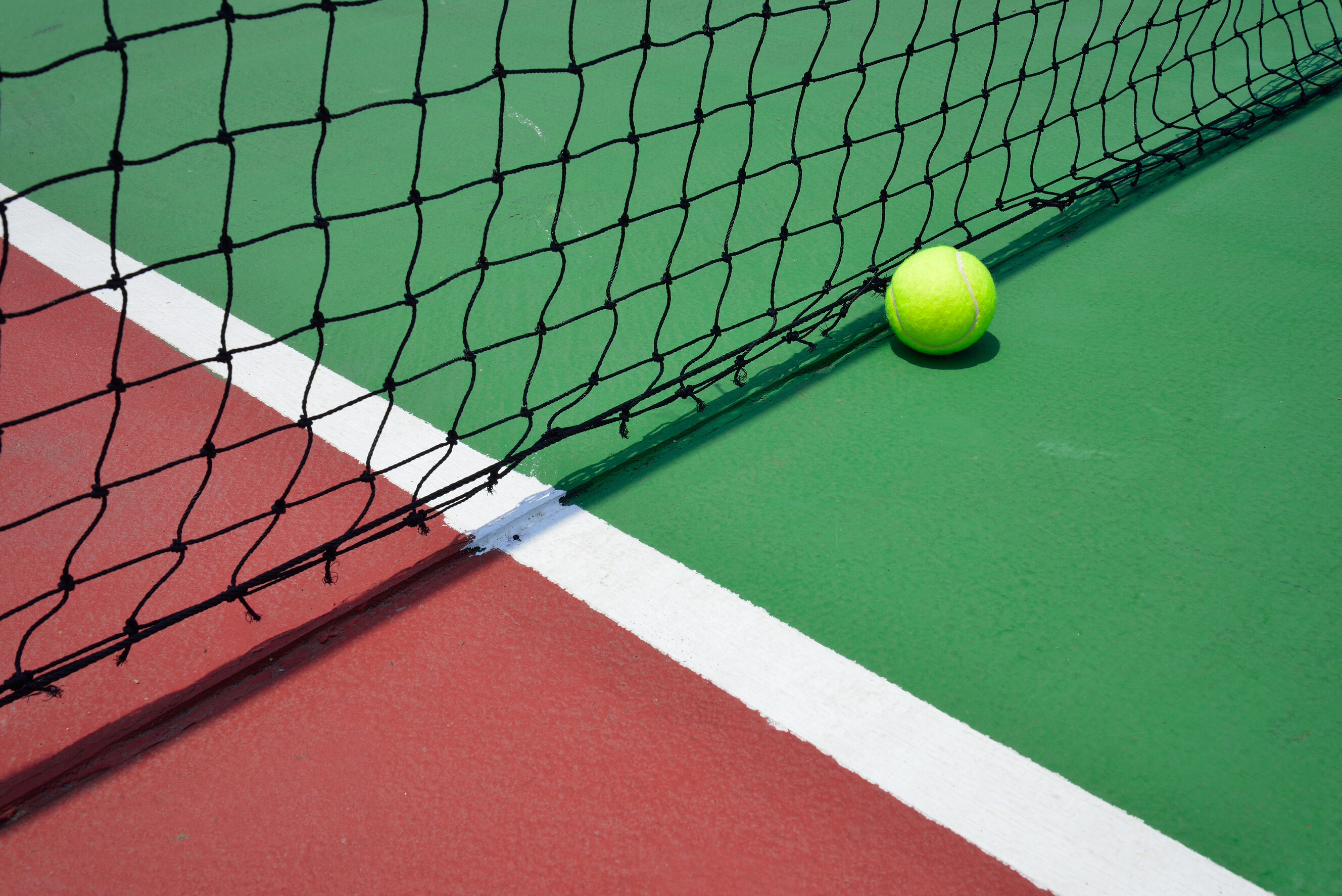 Tennis ball resting against the net on a two-tone acrylic hard court with white line markings.