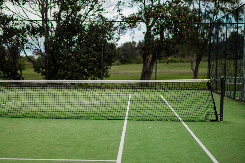 Ground-level view of a synthetic grass tennis court with net and tree-lined fencing in the background. - tennis court resurfacing cost