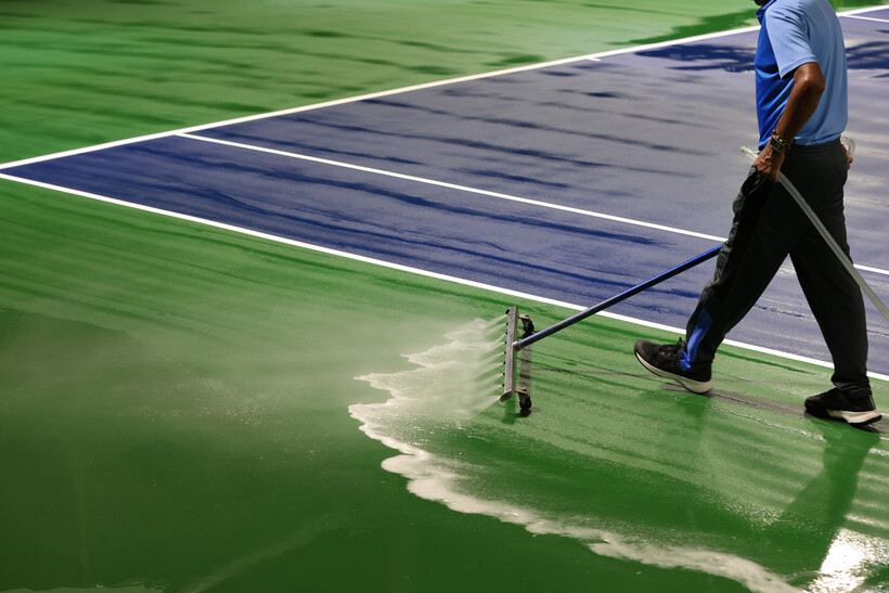 Court maintenance worker pressure washing a green acrylic tennis court surface with a long-handled squeegee. - tennis court resurfacing cost