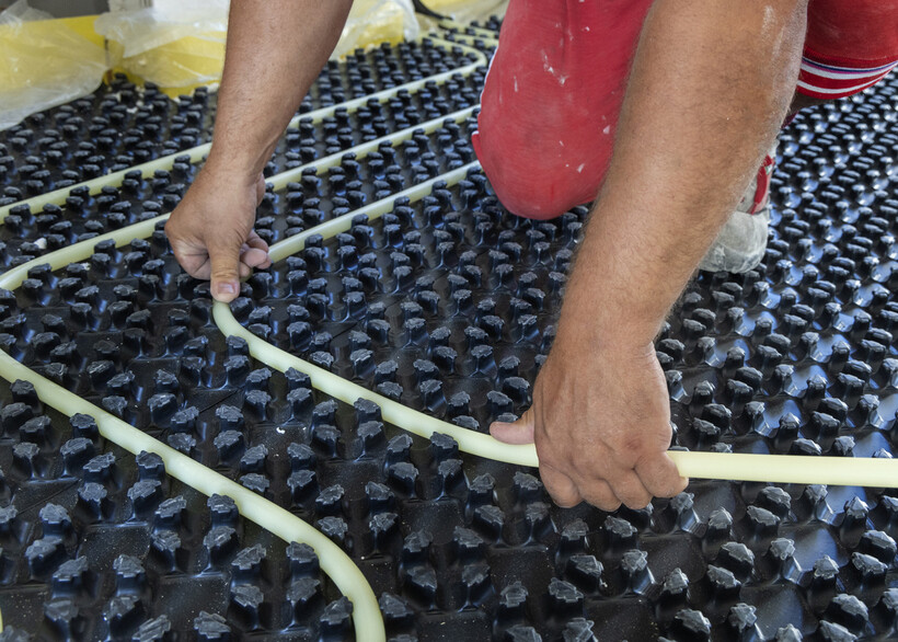 Close-up of hands guiding flexible pipe into studded subfloor insulation during installation. - underfloor heating installation cost