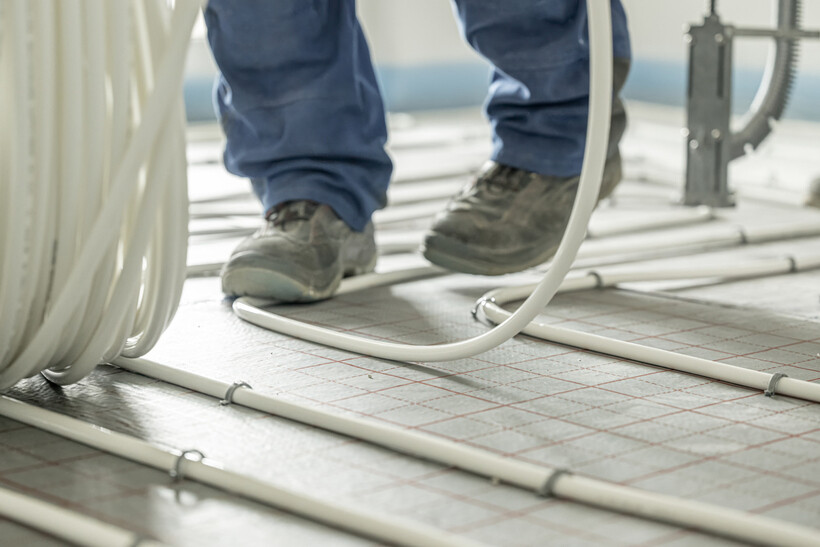 A worker’s boots stepping across a grid of white heating pipe on reflective underlay. - underfloor heating installation cost