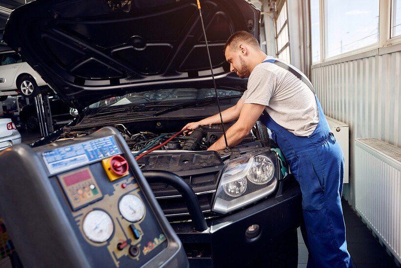 A mechanic works on a car's engine with air conditioning equipment nearby. - car aircon regas cost