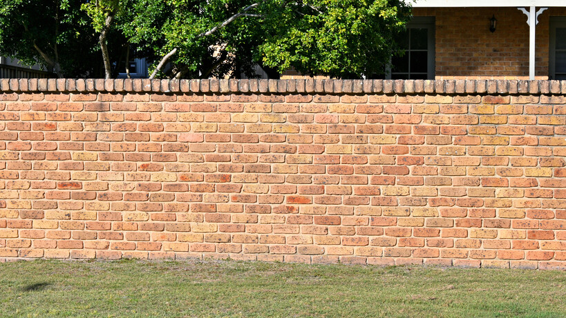 A tan brick fence with a capped top borders a grassed garden area. - brick fencing cost