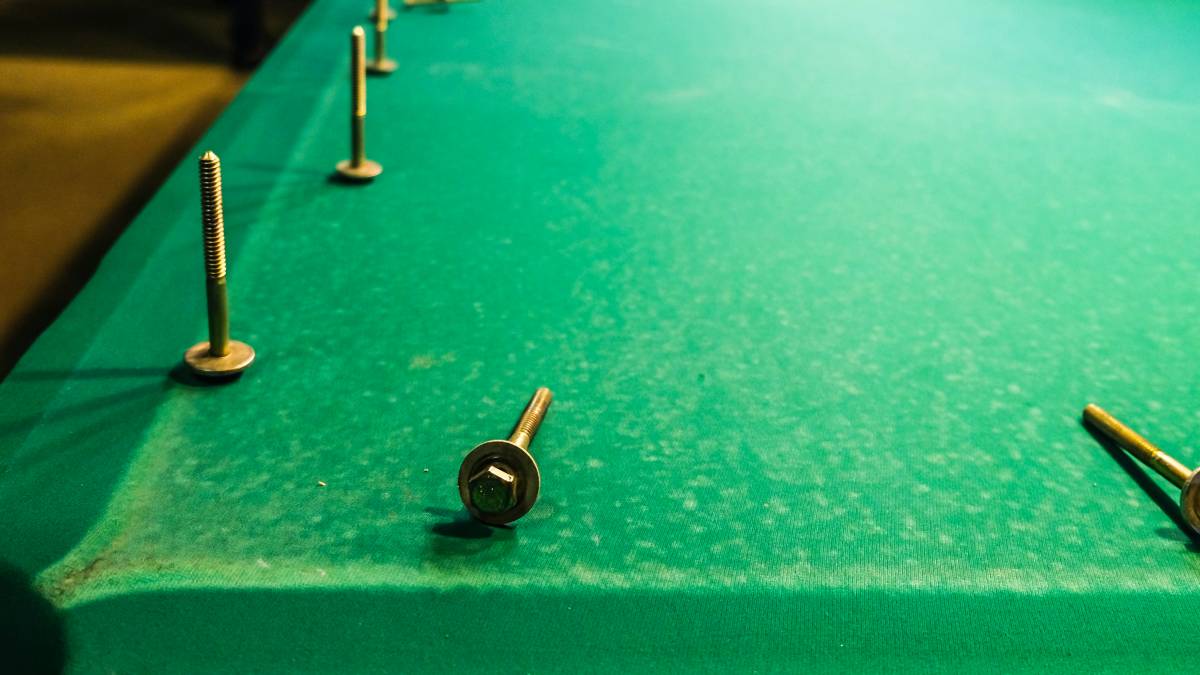 A close-up of a pool table undergoing refelting, with bolts removed and green felt partially worn.