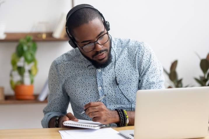 Focused man in headphones and watching online lesson on laptop, taking down notes from event management course