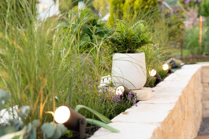 Garden illuminated with small spotlights, highlighting greenery and a white planter with ferns, placed on a stone ledge