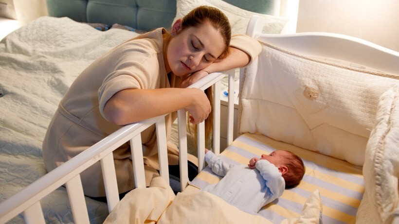Bassinet vs cot - A mother falling asleep while rocking the cot of her newborn baby at night