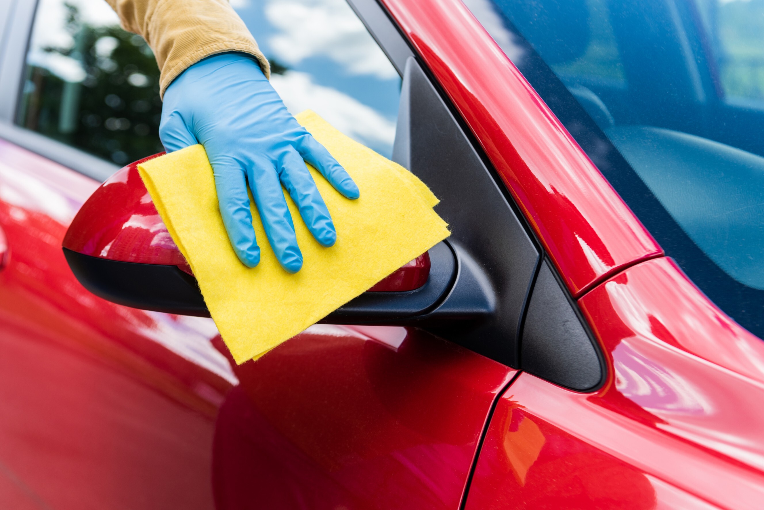 Close-up of a person cleaning a red car side mirror with a yellow cloth while wearing gloves. Discover car cleaning near me for professional services.