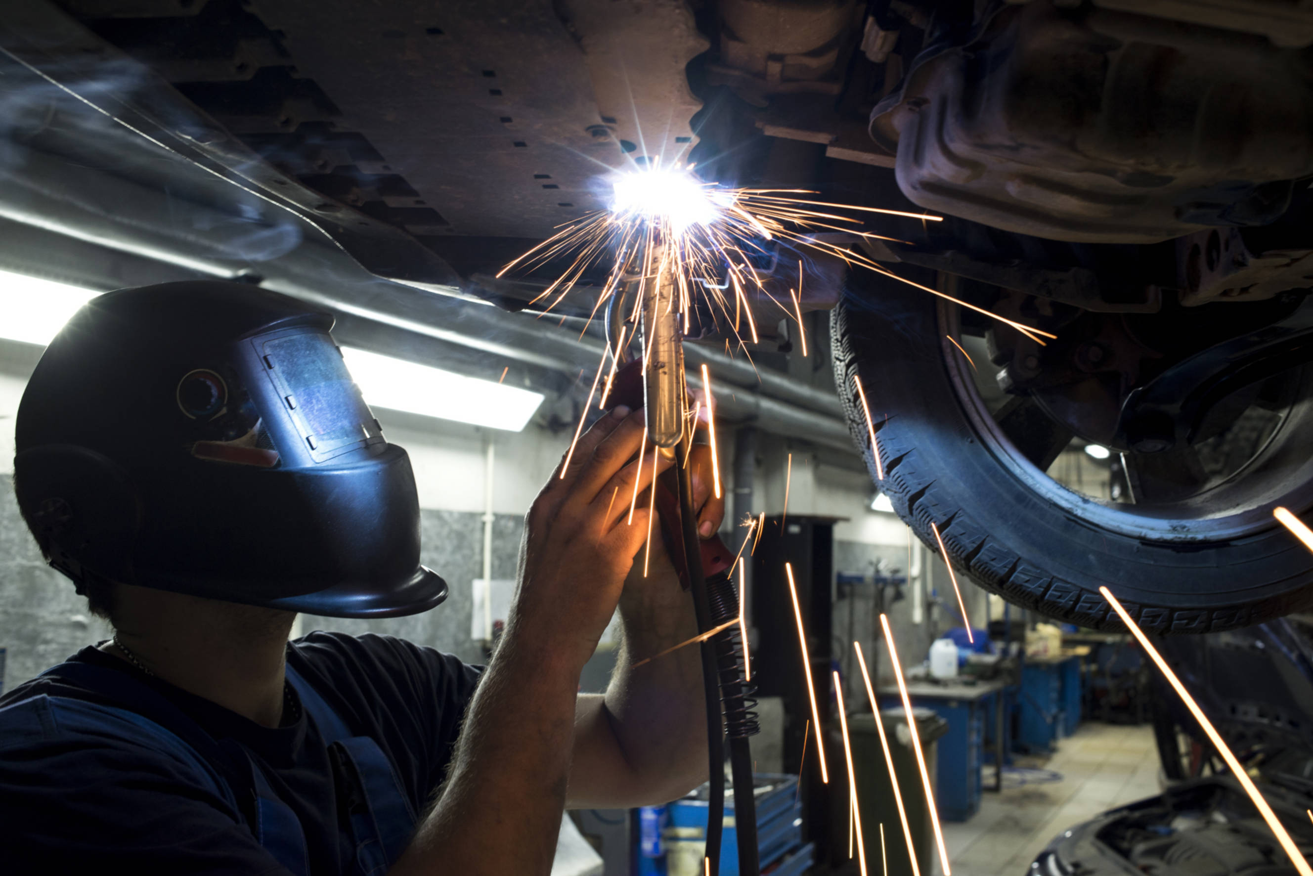 Technician performing car welding, using a welding tool to repair and strengthen the undercarriage of a vehicle in an auto workshop.