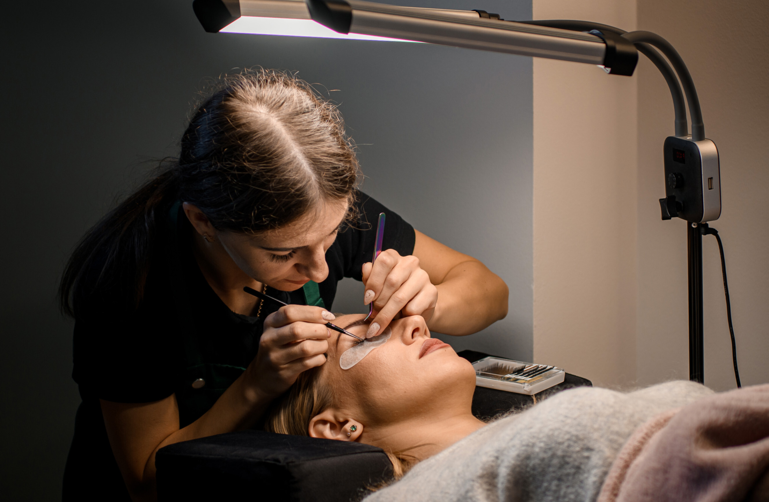 Beauty technician performing an eyelash lift and tint procedure on a client lying down.