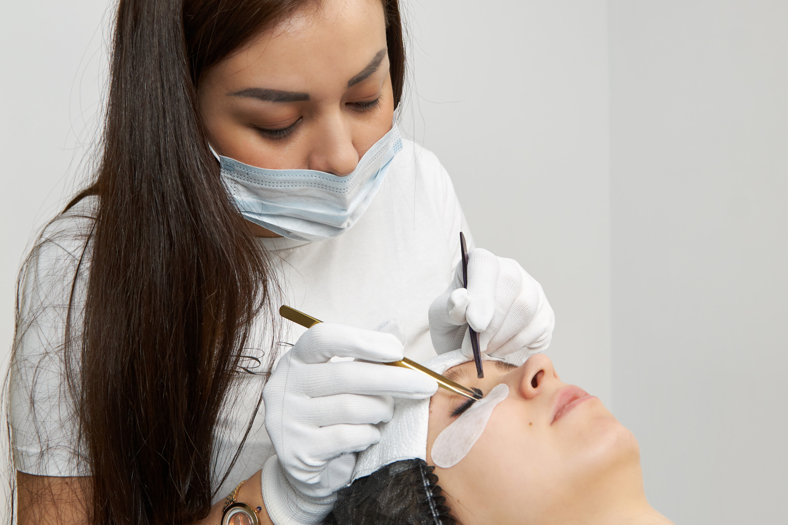 Beauty technician wearing gloves and a mask applying eyelash extensions to a client lying down.