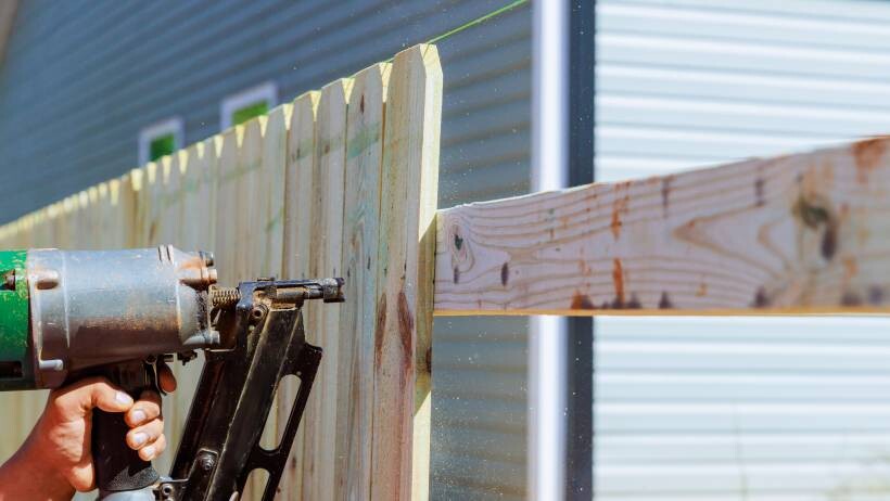 A person's hand operating a power tool to attach wooden boards to a fence in an outdoor setting - fencing cost