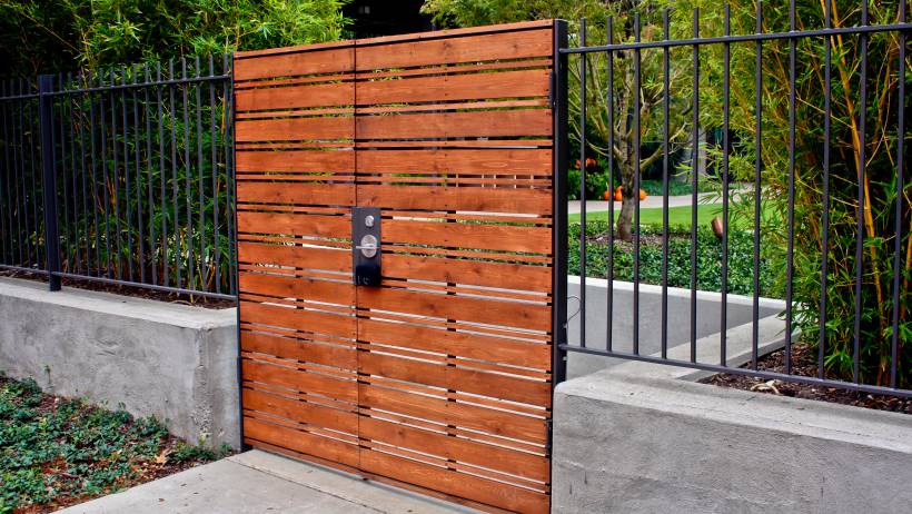 A wooden gate with horizontal slats is integrated into a black metal fence, surrounded by lush greenery in a landscaped garden setting. - fencing cost