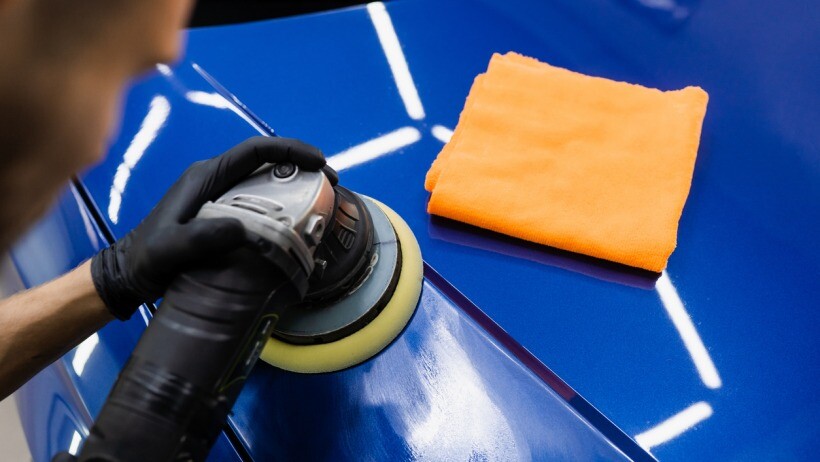 Car polish vs wax - A person using a power polishing tool to buff the surface of a blue car, with an orange microfiber cloth visible in the foreground.