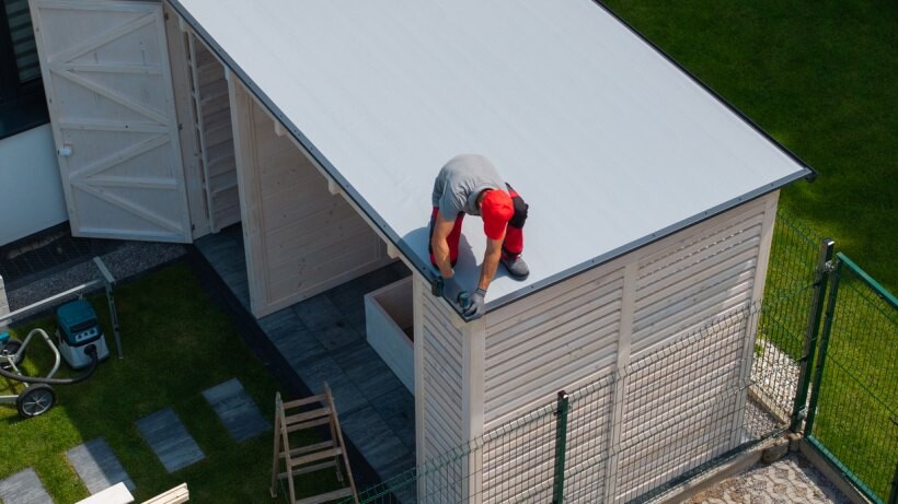 Person working on the roof of a white flat-roof shed in a neatly landscaped garden