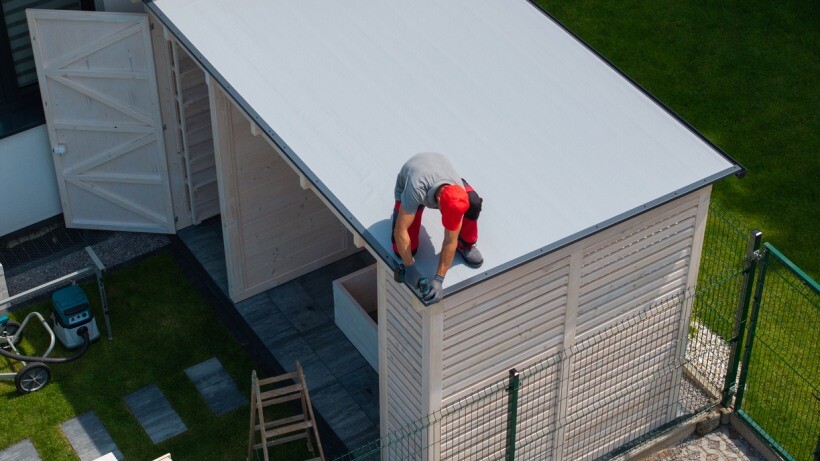 Barn vs shed - Person working on the roof of a small white shed with open doors, located in a fenced yard