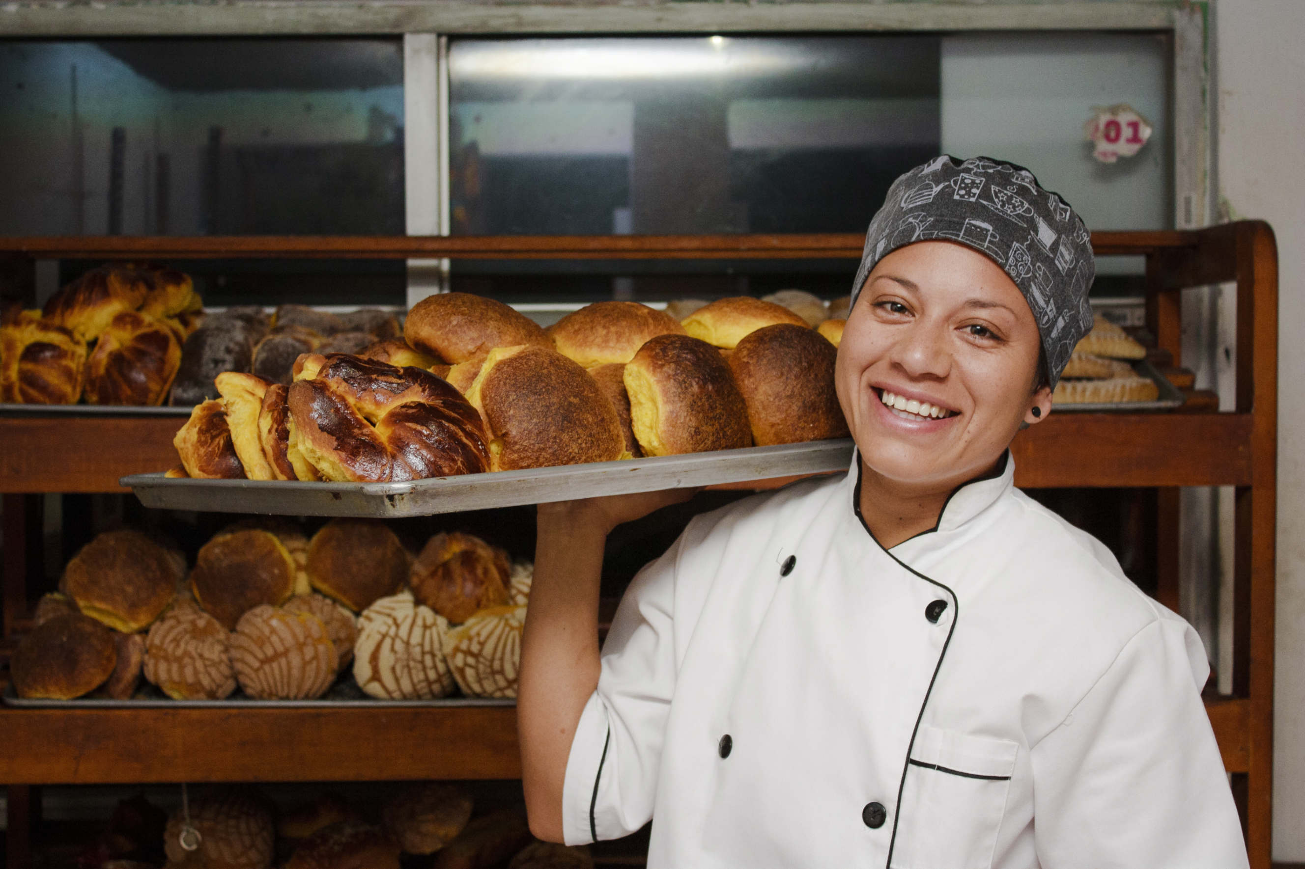 A smiling baker holding a tray of freshly baked bread in a bakery.