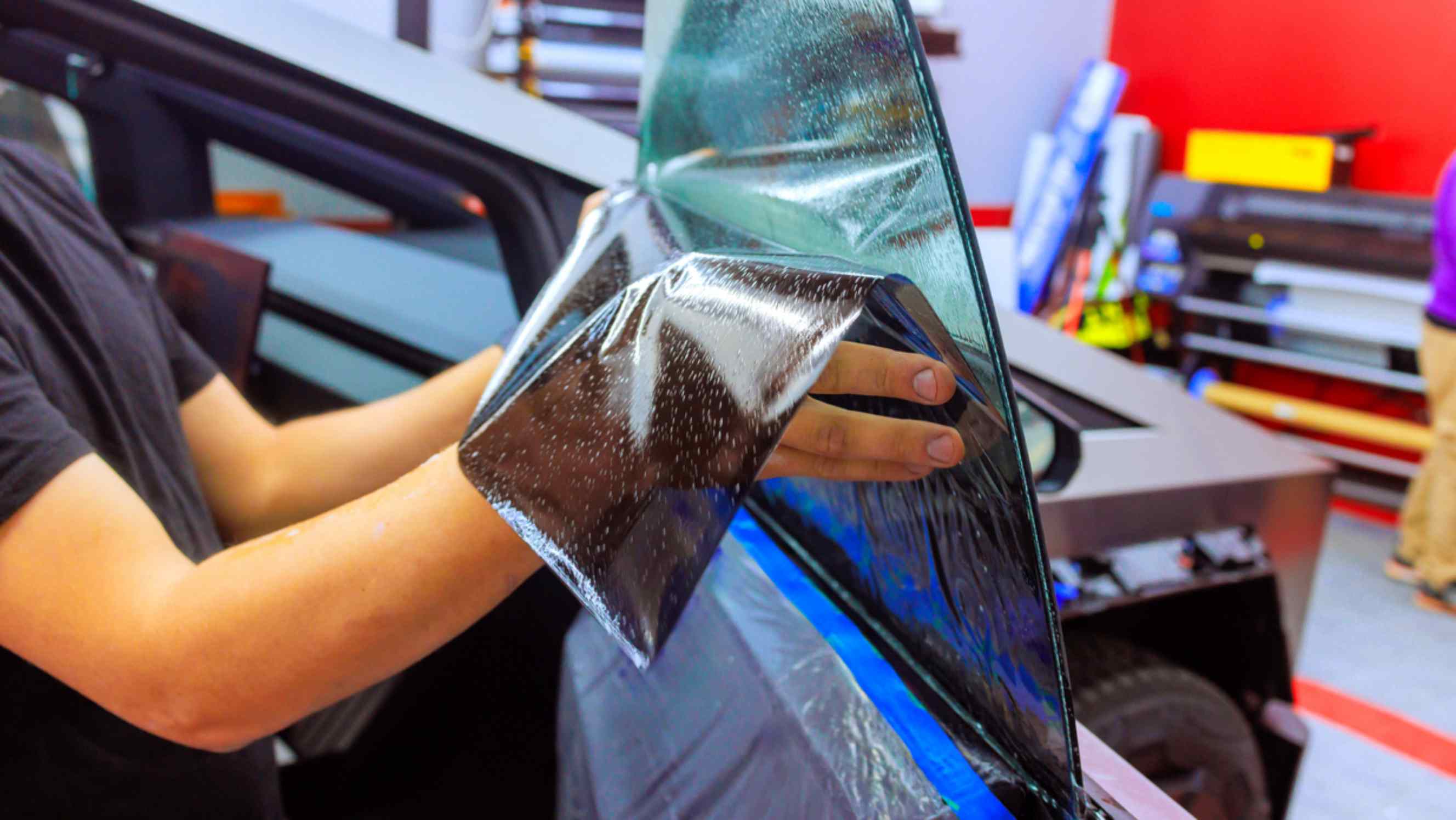 A technician carefully applying a tinted film to a car window in a workshop, representing professional car window tinting services near you.