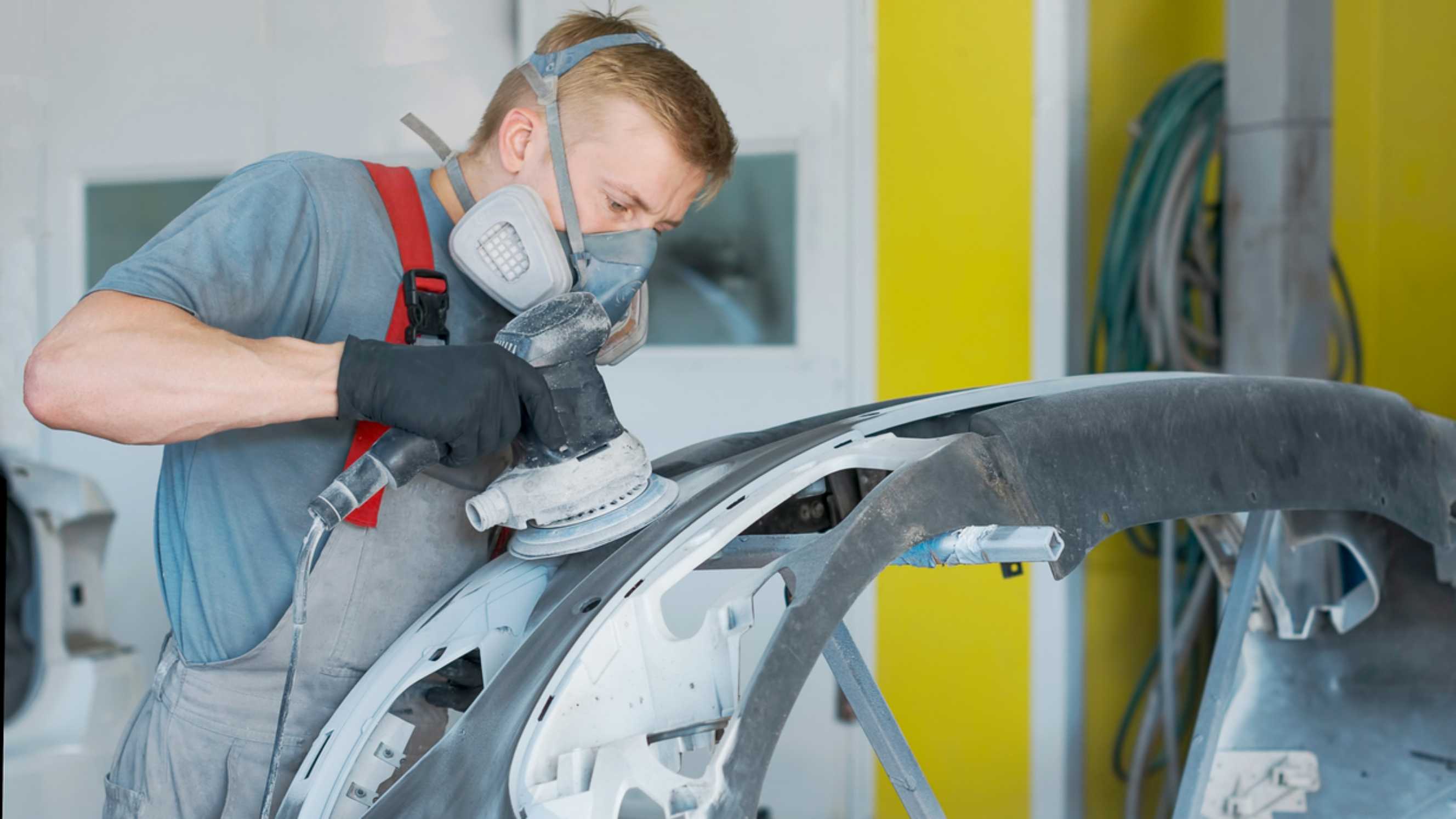 A technician wearing gloves and a mask is performing car body repair near you, using a power sander to smooth the surface of a car's bumper or body panel.