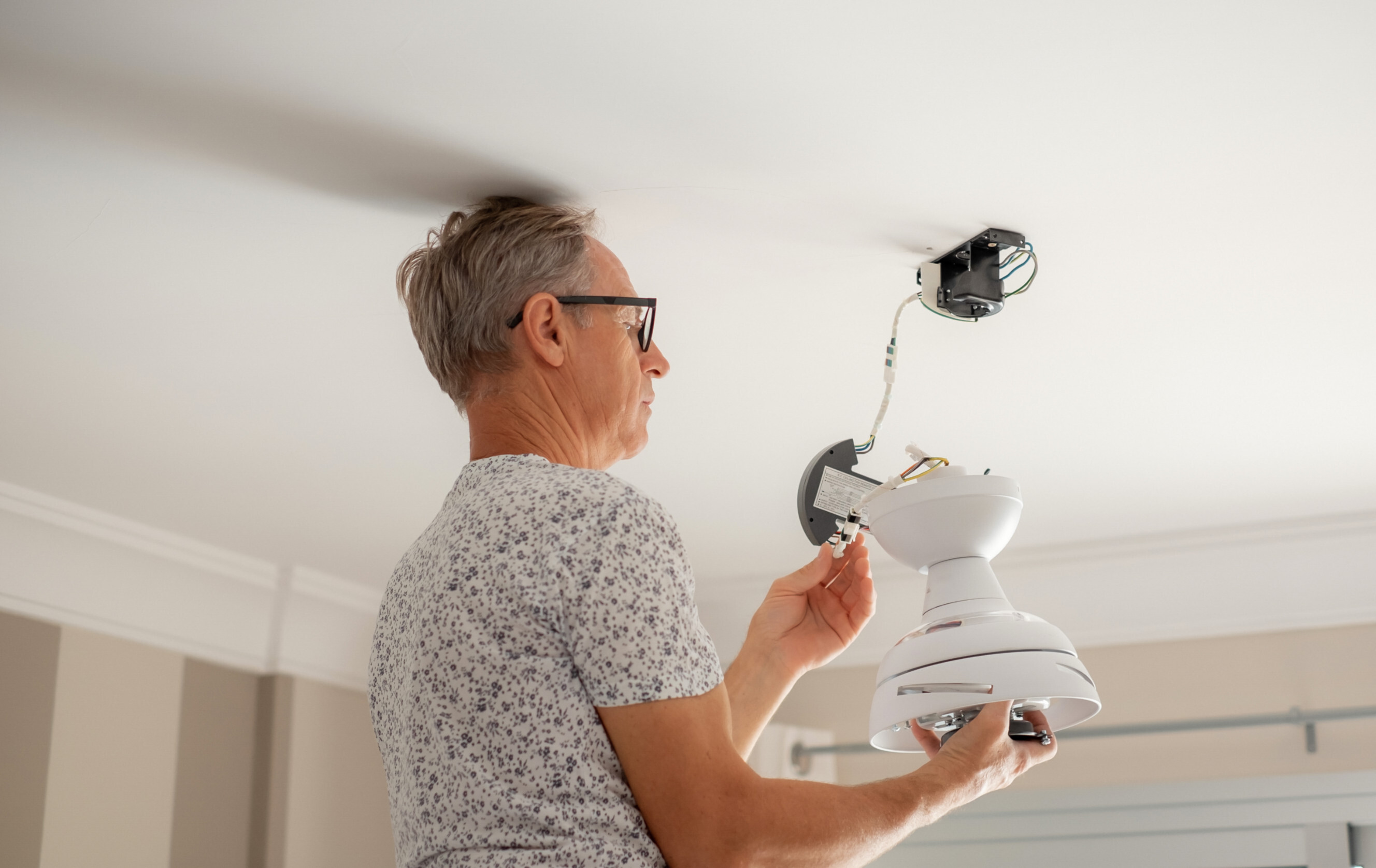 An elderly man installing a white ceiling fan in a modern home.