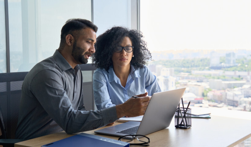 advisory-vs-consulting – two colleagues having a discussion while working on a laptop by a high-rise office window