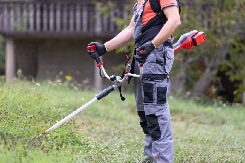 grass trimmer vs lawn mower - Person wearing protective gear while cutting grass with a battery-powered trimmer