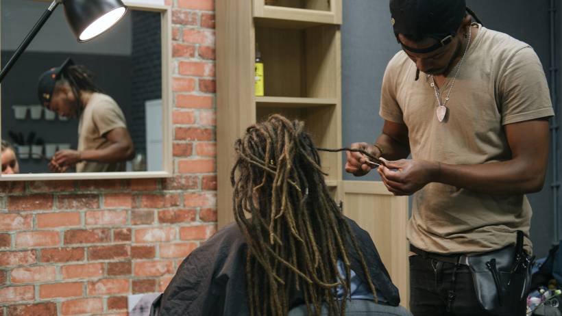 A male stylist in a tan shirt and black cap installing dreadlocks on a client in a salon with a brick wall and mirror. - dreadlocks cost