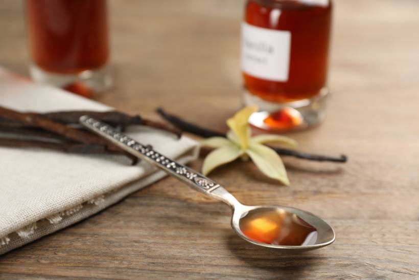 vanilla essence vs. extract - Vanilla extract on a spoon with vanilla beans and a vanilla flower on a wooden surface